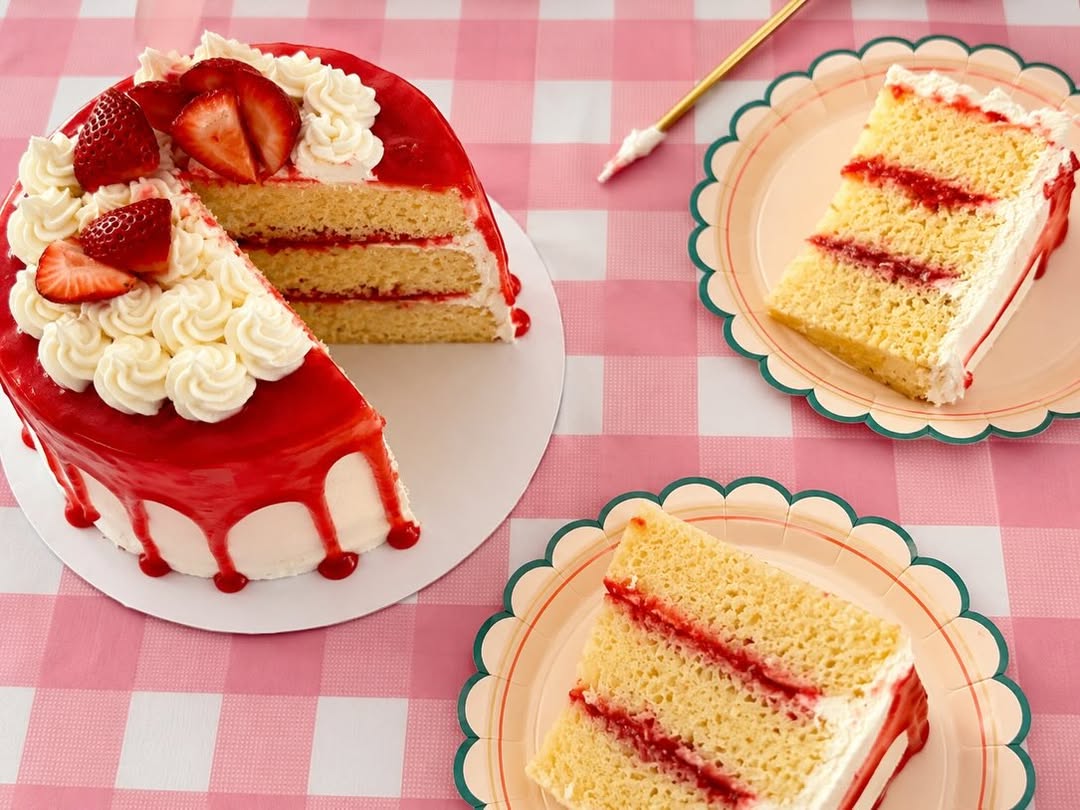 A strawberry layer cake with red glaze and fresh strawberries on top, two slices on pink scalloped plates.